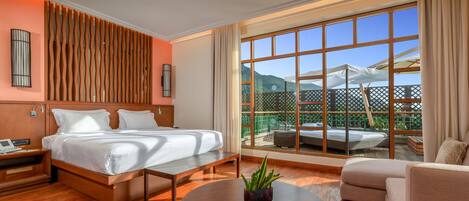 A modern hotel room with a large bed, a seating area, a round coffee table, and a view of a balcony with outdoor furniture.