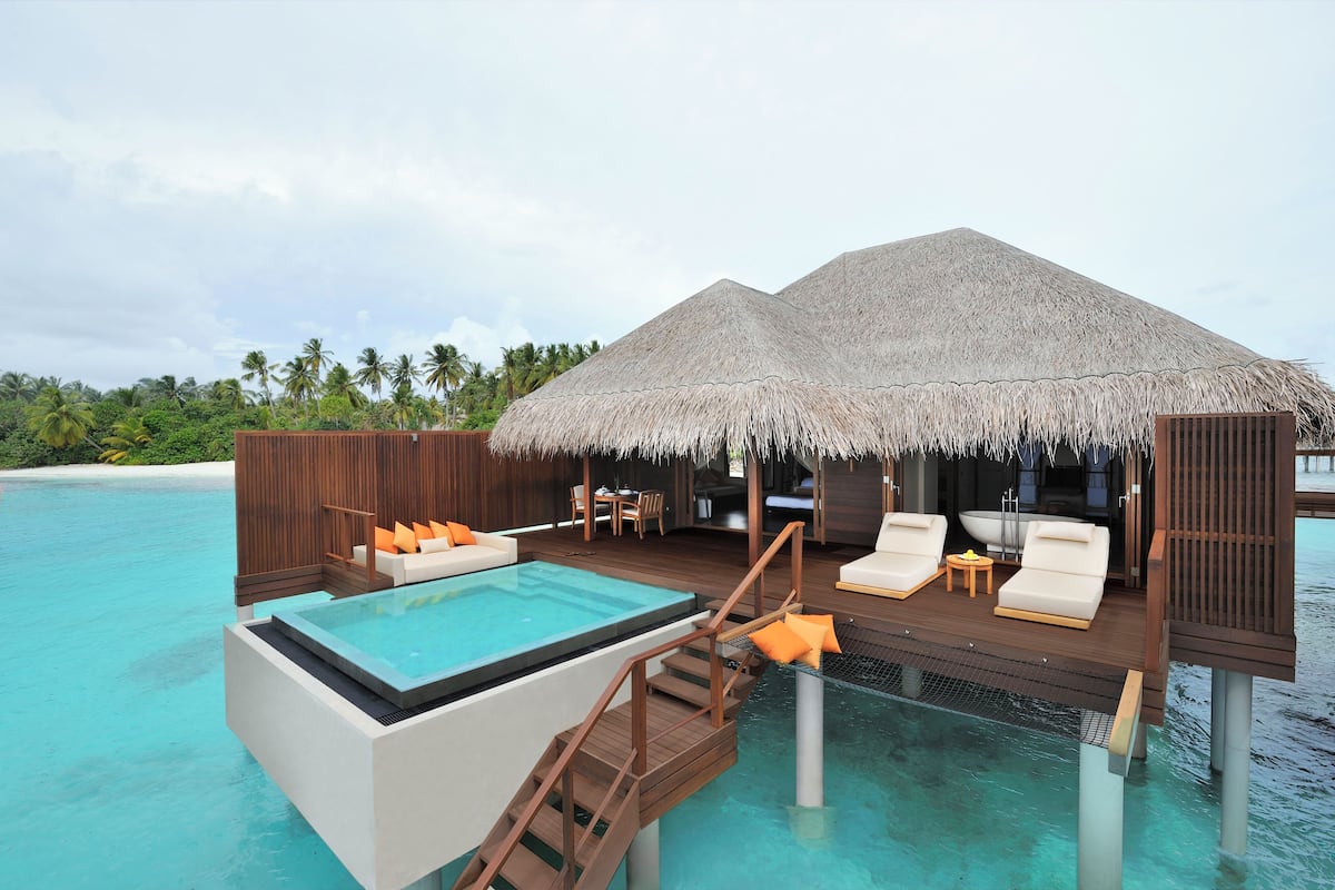 An overwater bungalows with a pool, lounge chairs, and a thatched roof.