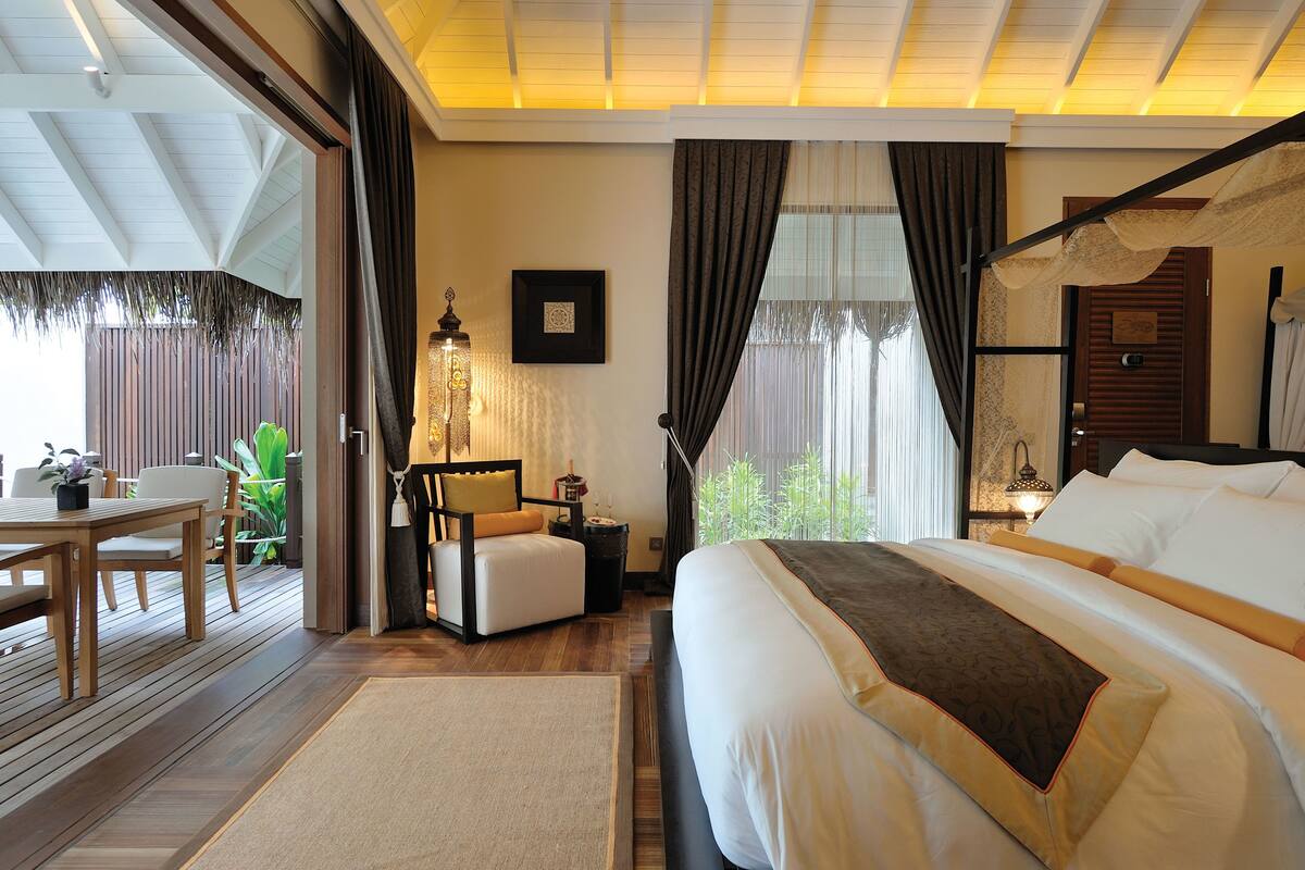 A hotel room with a large bed, a wooden desk, a chair, a small table, and a view of greenery outside.