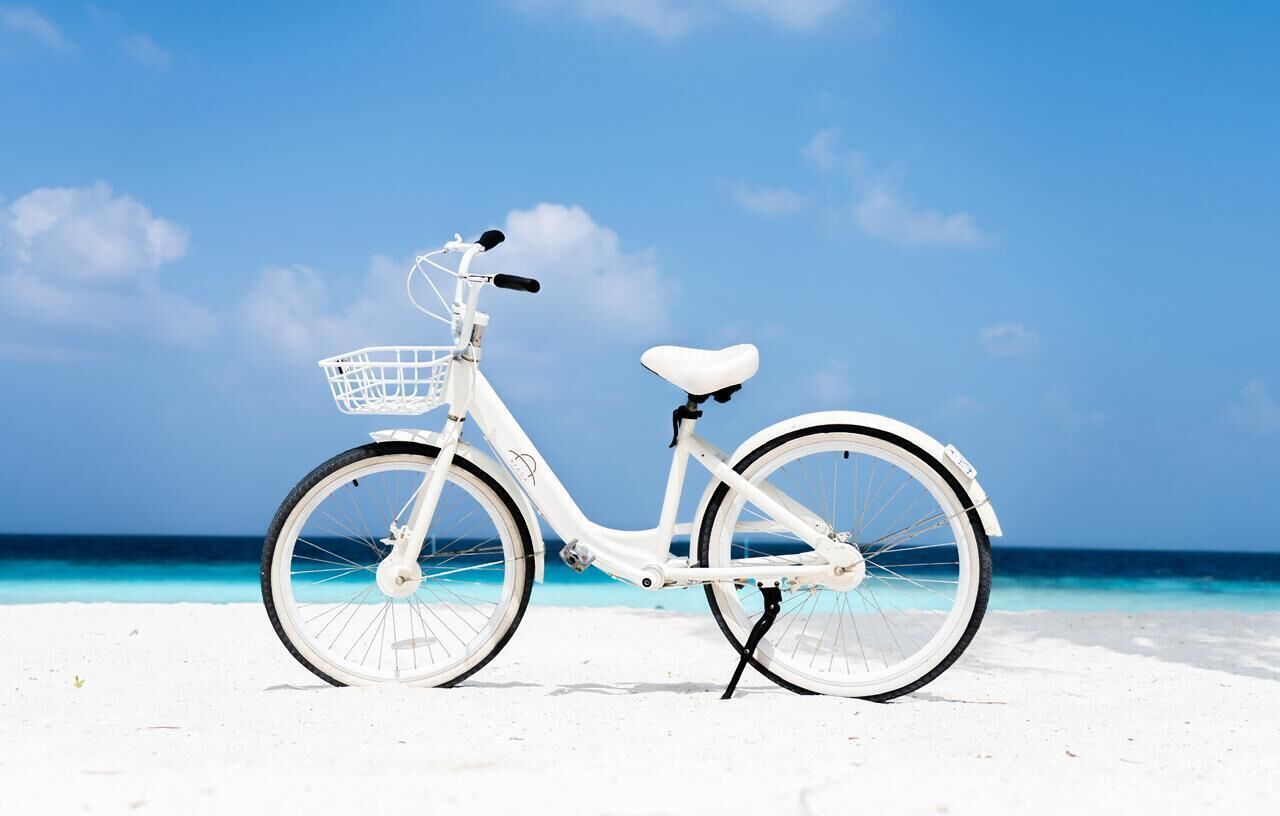 Ein weißes Fahrrad mit Korb am sandigen Strand, im Hintergrund klares, blaues Wasser.