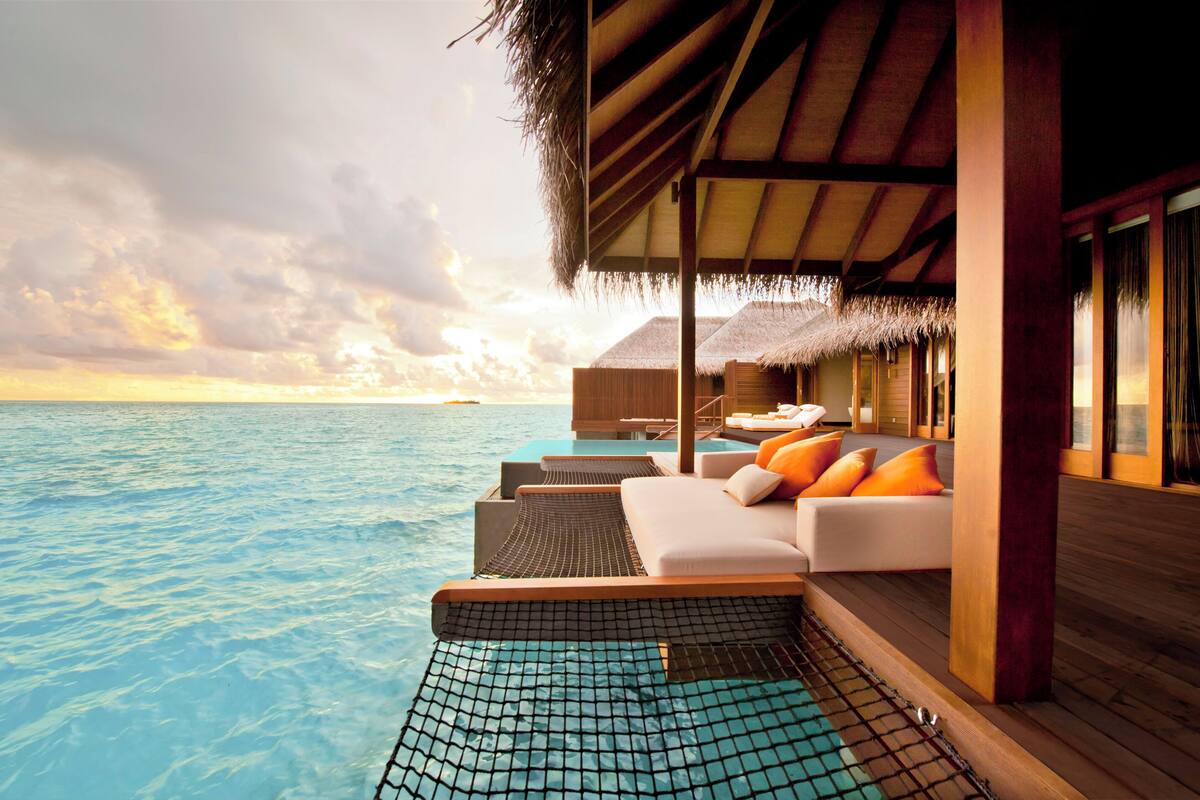 An overwater bungalows with a thatched roof, a pool, and a lounge area with cushions.