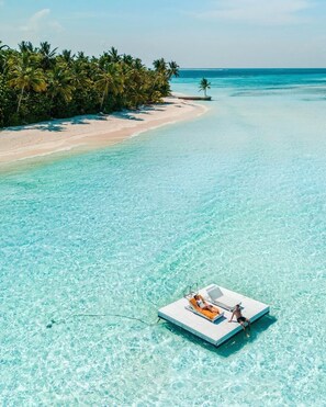 A clear turquoise sea with a sandy beach and palm trees. A small boat with two people is anchored near the shore. - Ayada Maldives (Maguhdhuvaa)