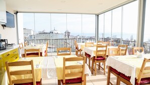 Comedor con varias mesas y sillas, ventanas amplias que ofrecen vista a la ciudad y una zona de cocina con armarios amarillos.