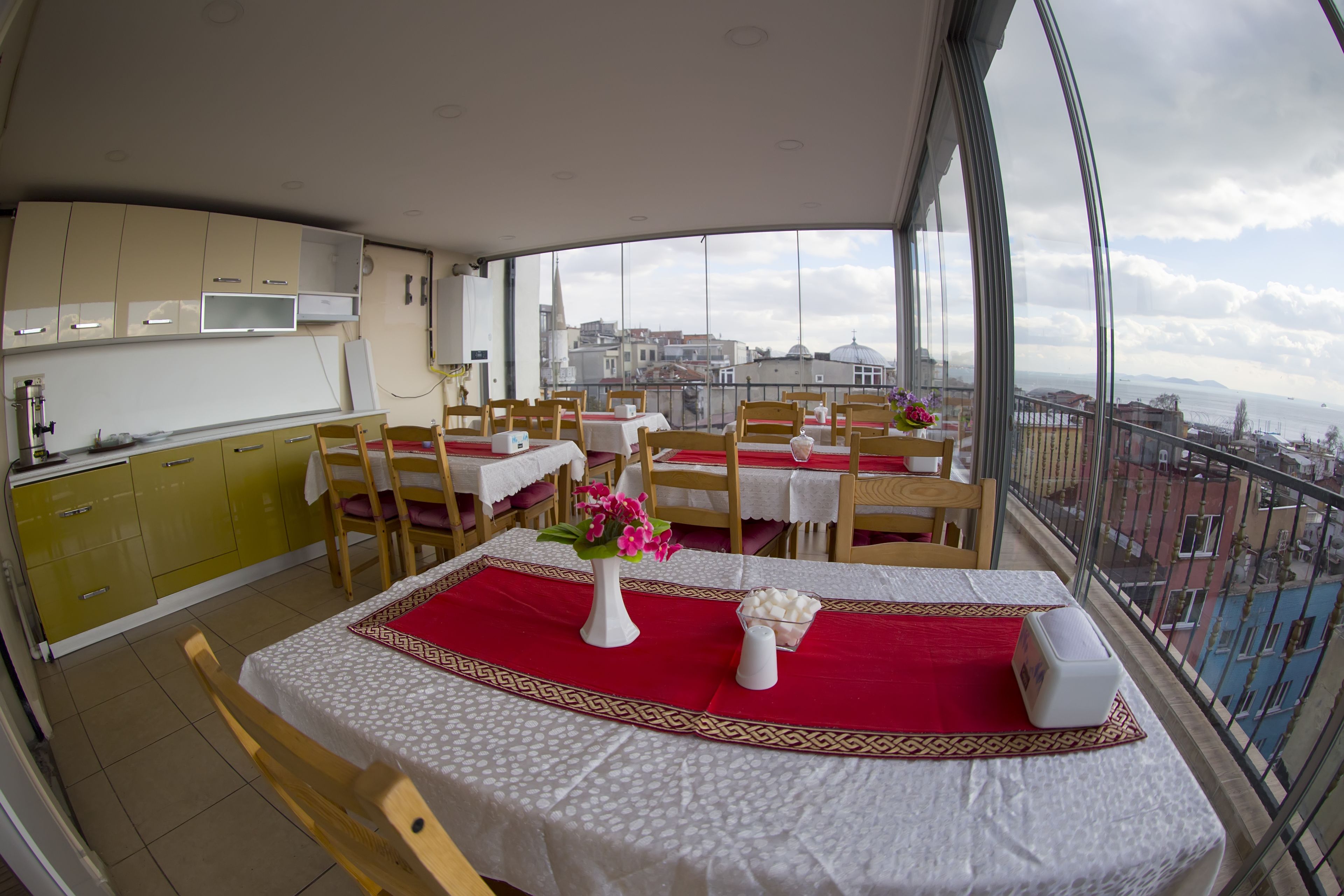 A dining area with tables set for two, a kitchen area with yellow cabinets, and a view of the city through large windows.