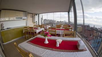 A dining area with tables set for two, a kitchen area with yellow cabinets, and a view of the city through large windows.