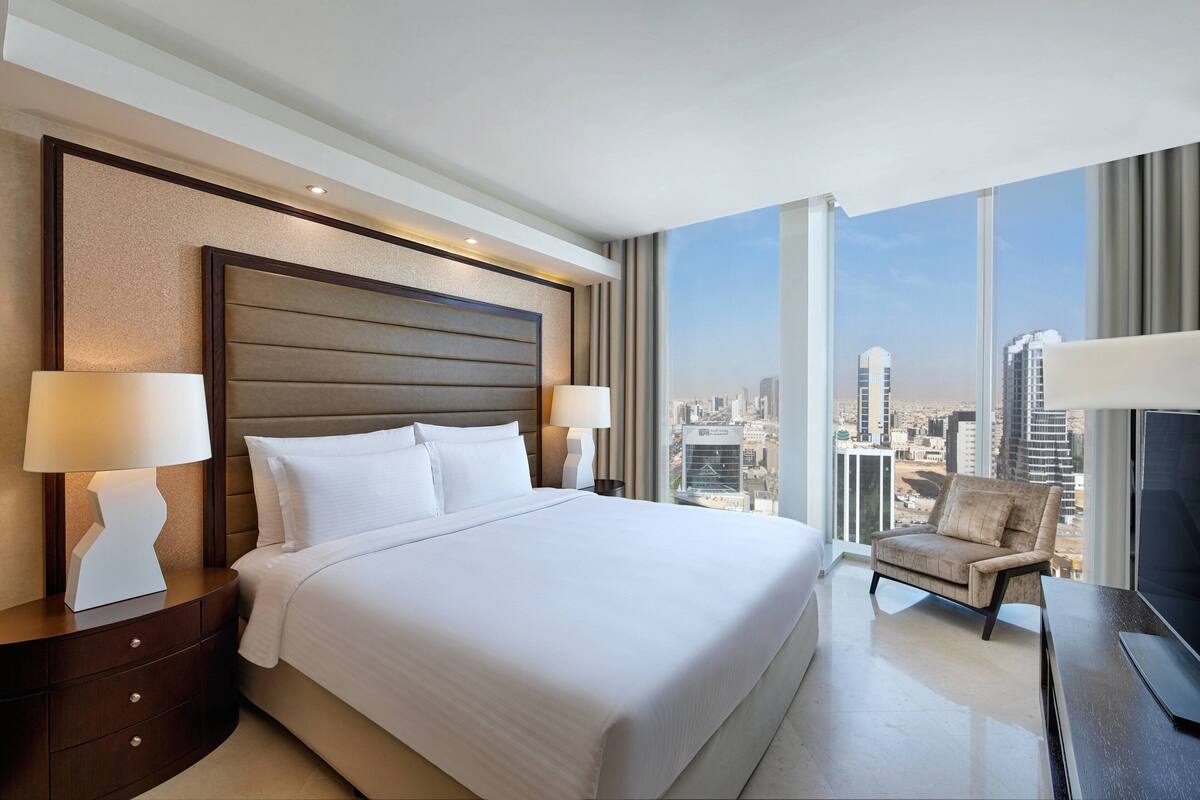 Una habitación de hotel moderna con una cama grande, dos lámparas de noche, una silla y un televisor. La habitación tiene vistas a la ciudad a través de amplias ventanas.