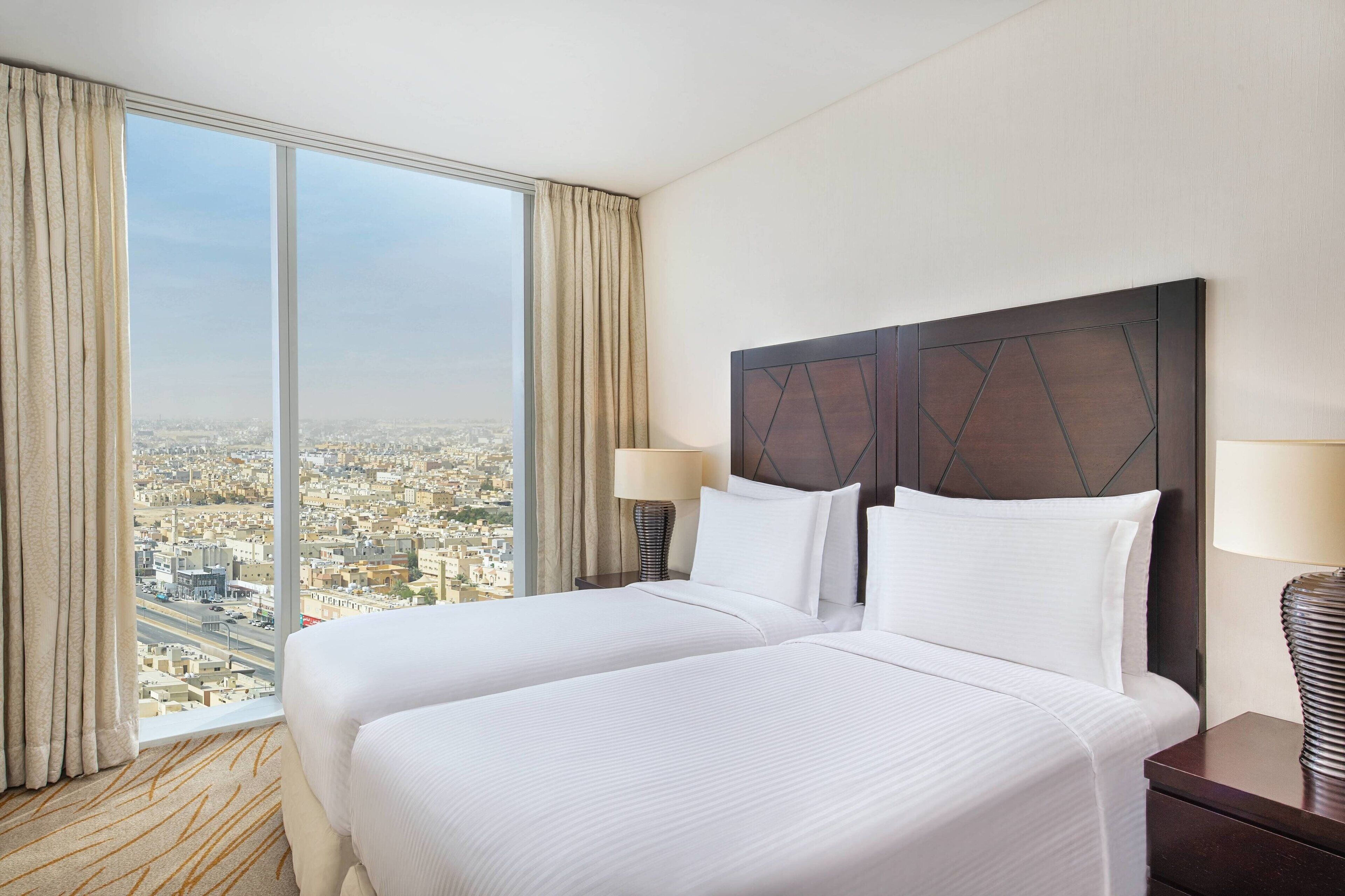 A hotel room with two beds, a large window with a city view, and a dark headboard.