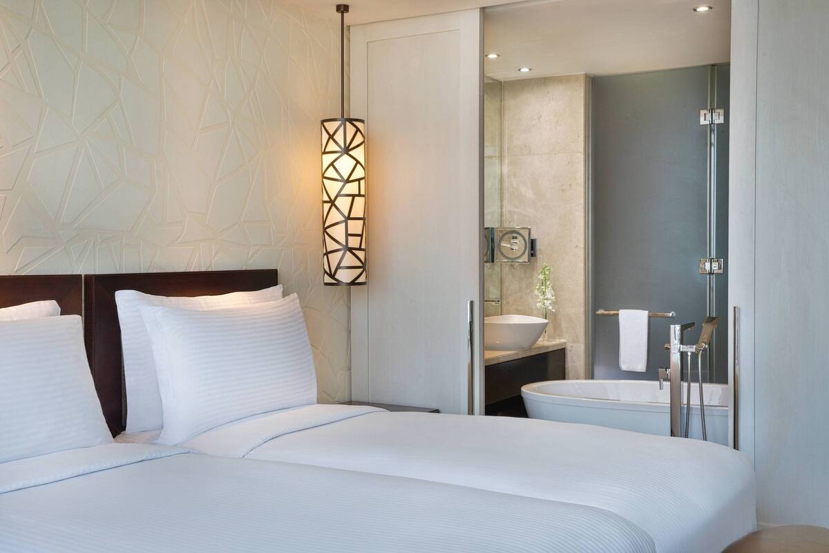 Una habitación de hotel moderna con una cama grande, una mesita de noche, una lámpara de pared, un baño con lavamanos y espejo, y una ducha.