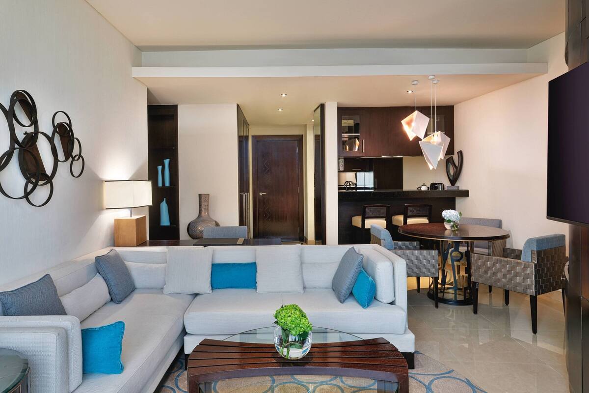 Una sala moderna con un sofá blanco, cojines azules, una mesa de centro de vidrio con una planta y una zona de comedor con una mesa redonda y sillas.