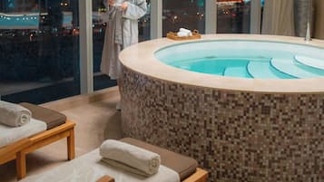A modern spa area with a mosaic-tiled hot tub, wooden loungers, and a view of the city at night.