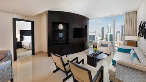 A modern hotel room with a curved black entertainment unit, a dining area with a glass table, a sofa, and a city view through large windows.