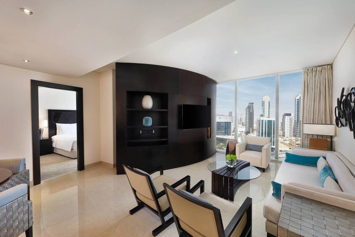 Una habitación de hotel moderna con un mueble de entretenimiento negro curvo, una zona de comedor con una mesa de vidrio, un sofá y vistas a la ciudad a través de amplios ventanales.