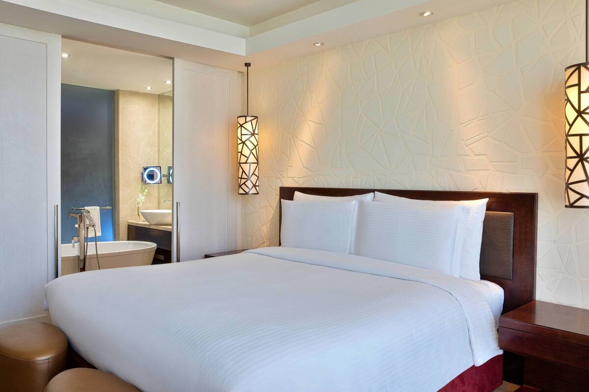Una habitación de hotel moderna con una cama grande, cabecera de madera y un baño visible a través de una puerta abierta.
