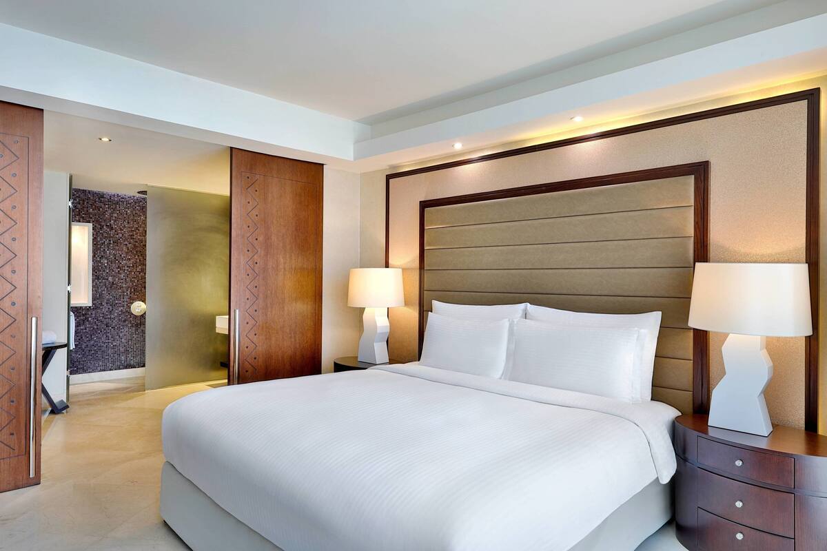 Una habitación de hotel moderna con una cama grande, dos mesitas de noche con lámparas, un cabecero de madera y un vestidor.