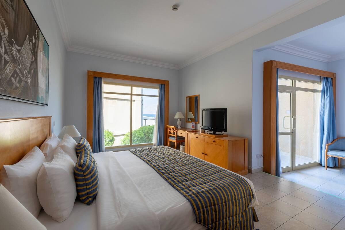 A hotel room with a large bed, a desk with a computer, a television, and a view of the outdoors through a sliding glass door.