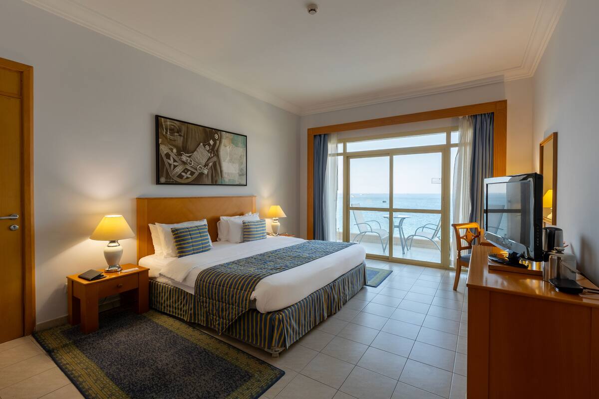 A hotel room with a large bed, a desk with a TV, a window with a view, and a painting on the wall.