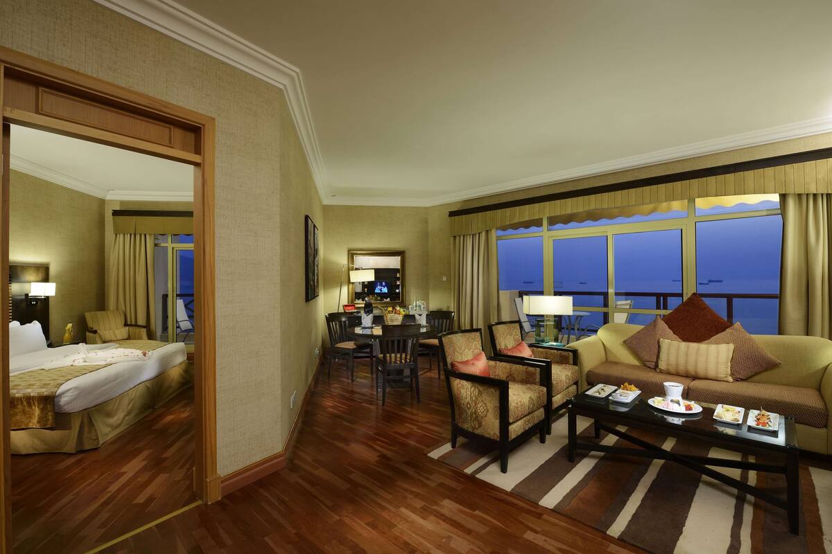 A hotel room with a bed, a sofa, a dining area, and a view of the city.