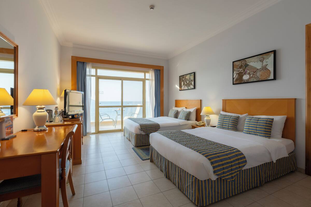 A hotel room with two beds, a desk, a chair, a TV, and a large window with a view.
