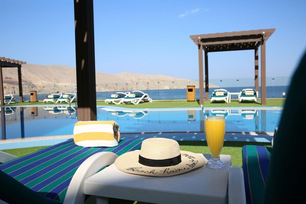 A poolside lounge area with a view of the sea, lounge chairs, and a table with a hat and a glass of orange juice.