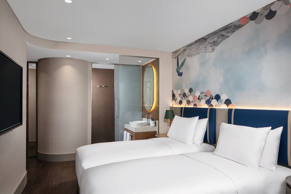 A modern hotel room with a large bed, a bathroom with a glass partition, and a wall mural.