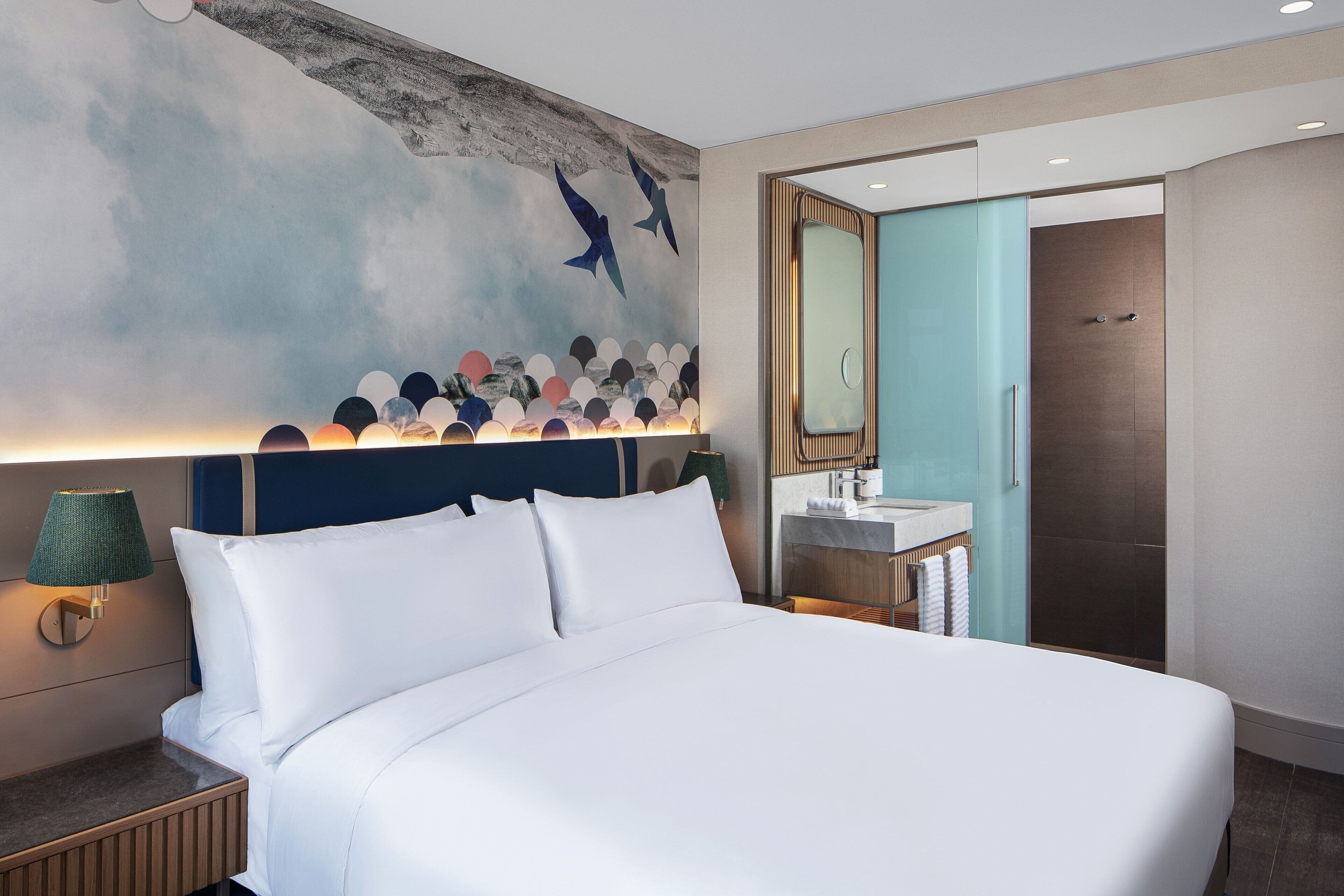 A modern hotel room with a large bed, a nightstand with a lamp, a bathroom with a sink and mirror, and a wall mural.