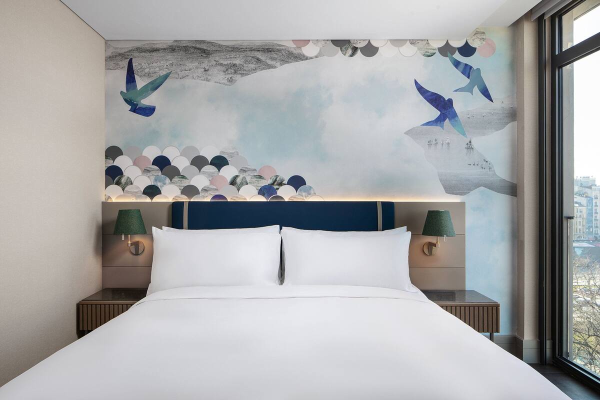 A hotel room with a large bed, two nightstands with lamps, a mural with birds and abstract shapes, and a view through a window.