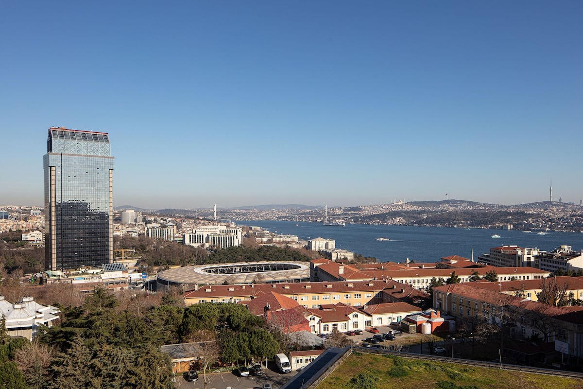 A high-rise building with a glass facade, surrounded by a mix of residential and commercial buildings, with a body of water and distant hills in the background.