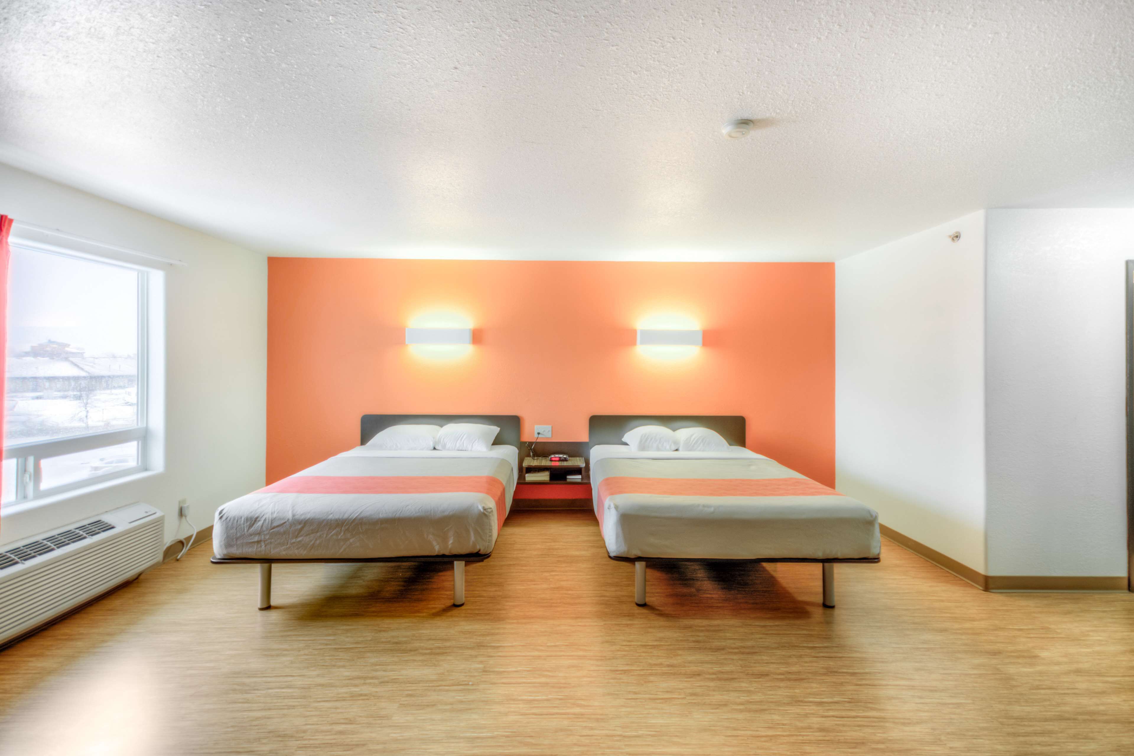 A hotel room with two beds, a window, an air conditioner, and a wall-mounted light fixture.