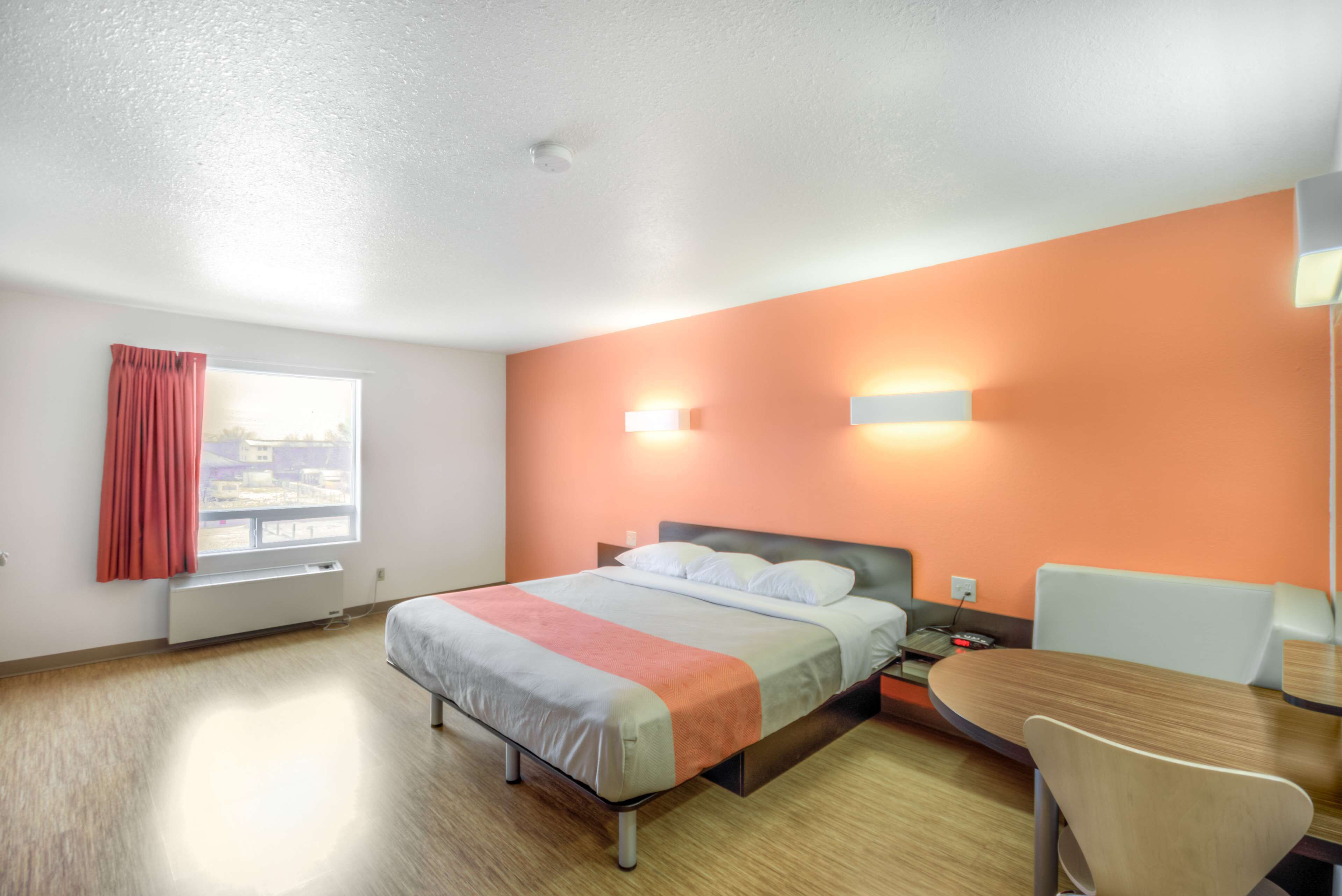 A hotel room with a bed, a desk, and a chair. There is a window with curtains, a radiator, and a wall-mounted light fixture.
