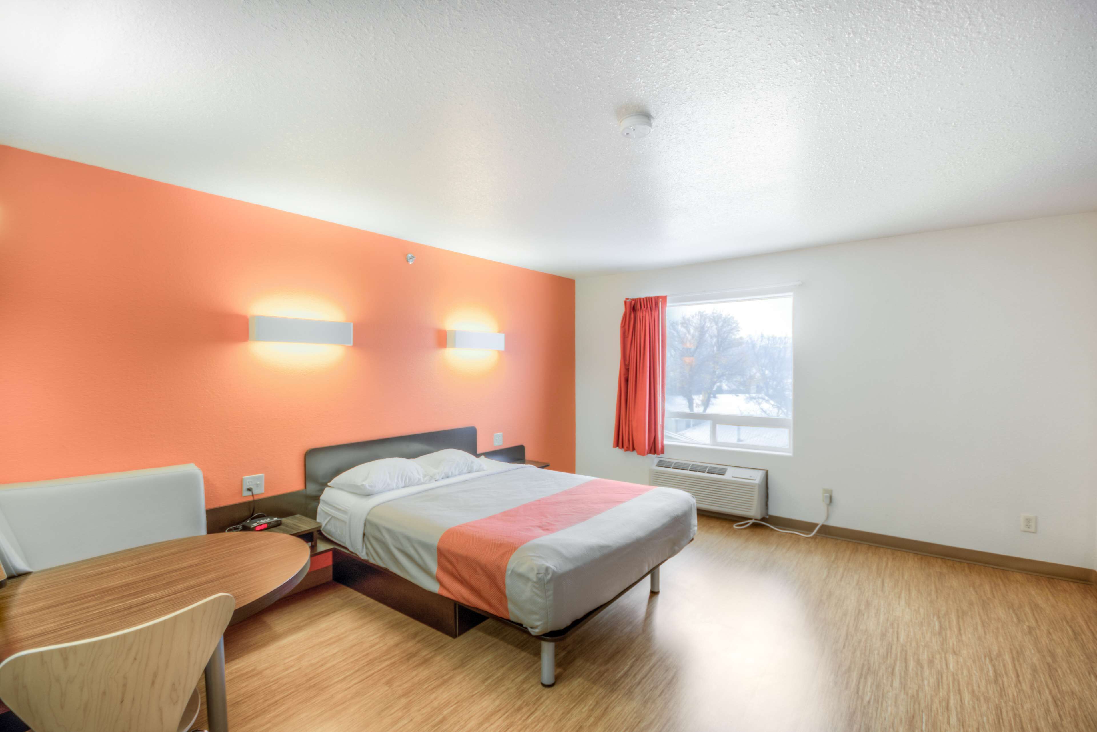 A hotel room with a bed, a desk, and a chair. There is a window with curtains and a view of trees outside.