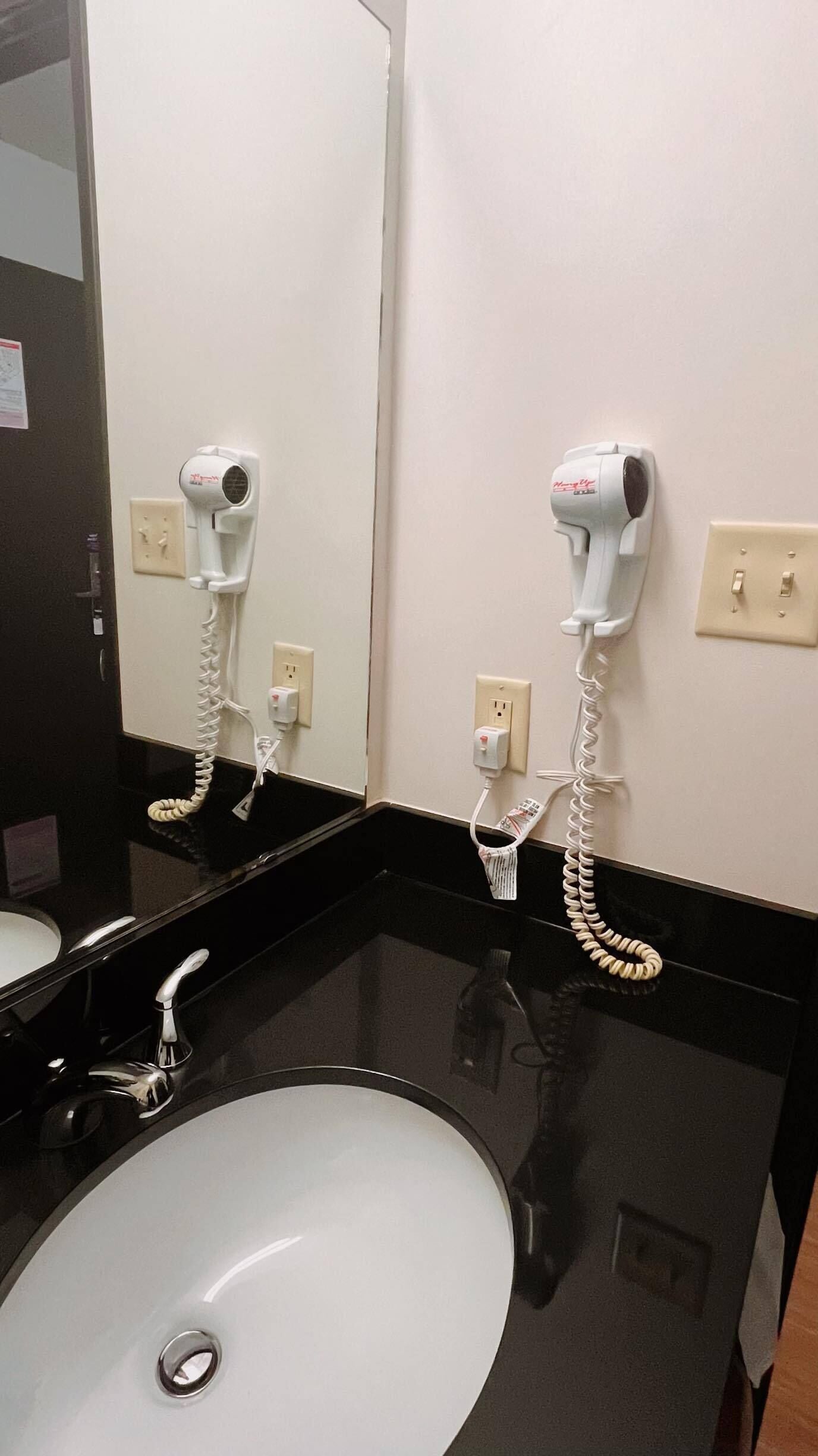 A hotel bathroom with a white sink, a mirror, and two hair dryers mounted on the wall.