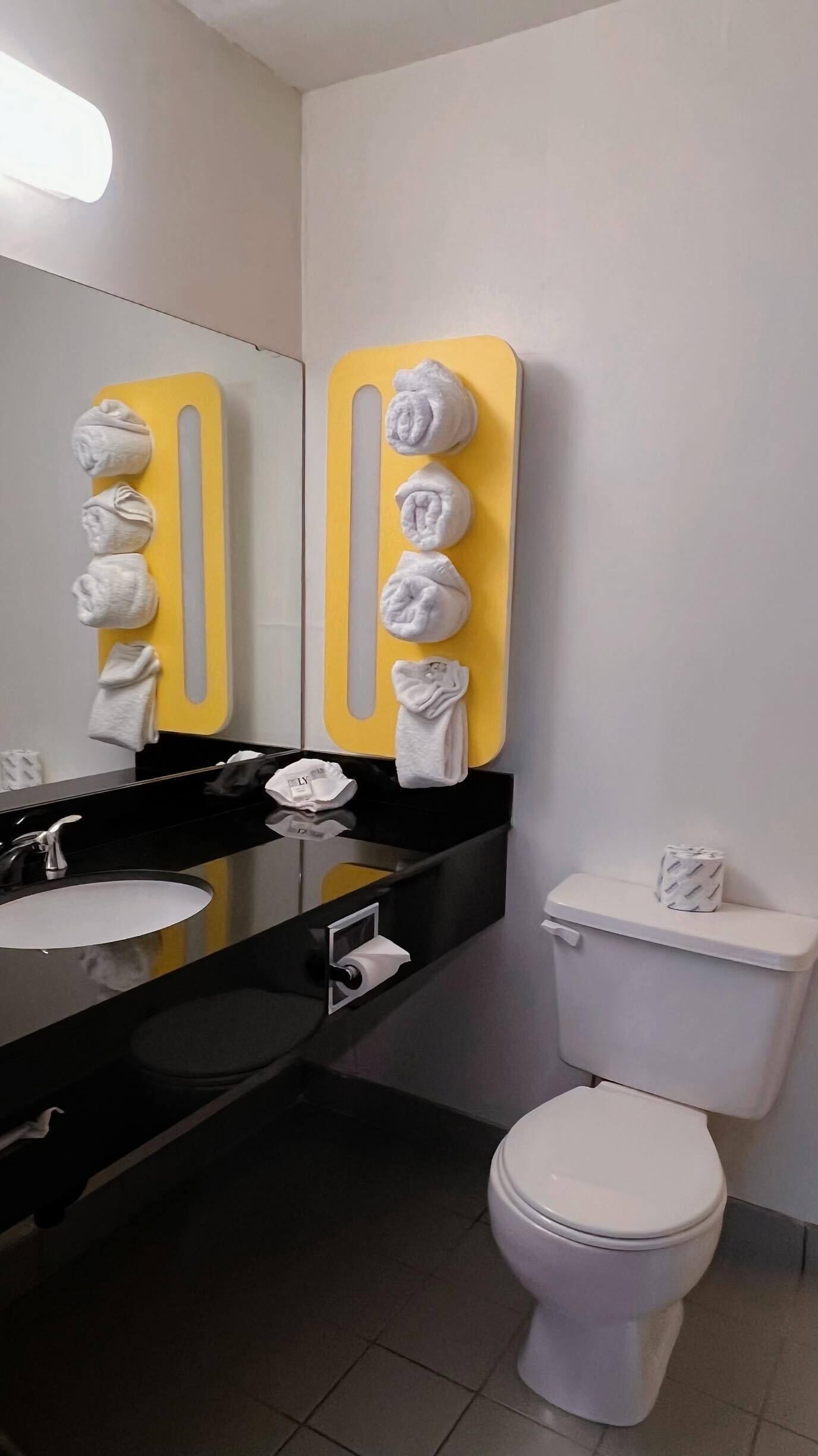 Un baño con un lavamanos negro, un espejo y un inodoro. Toallas cuelgan de un perchero amarillo.