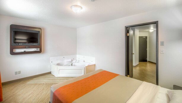 A hotel room with a bed, a large mirror, a television mounted on the wall, and a bathtub. - Motel 6 Regina, SK (Regina)