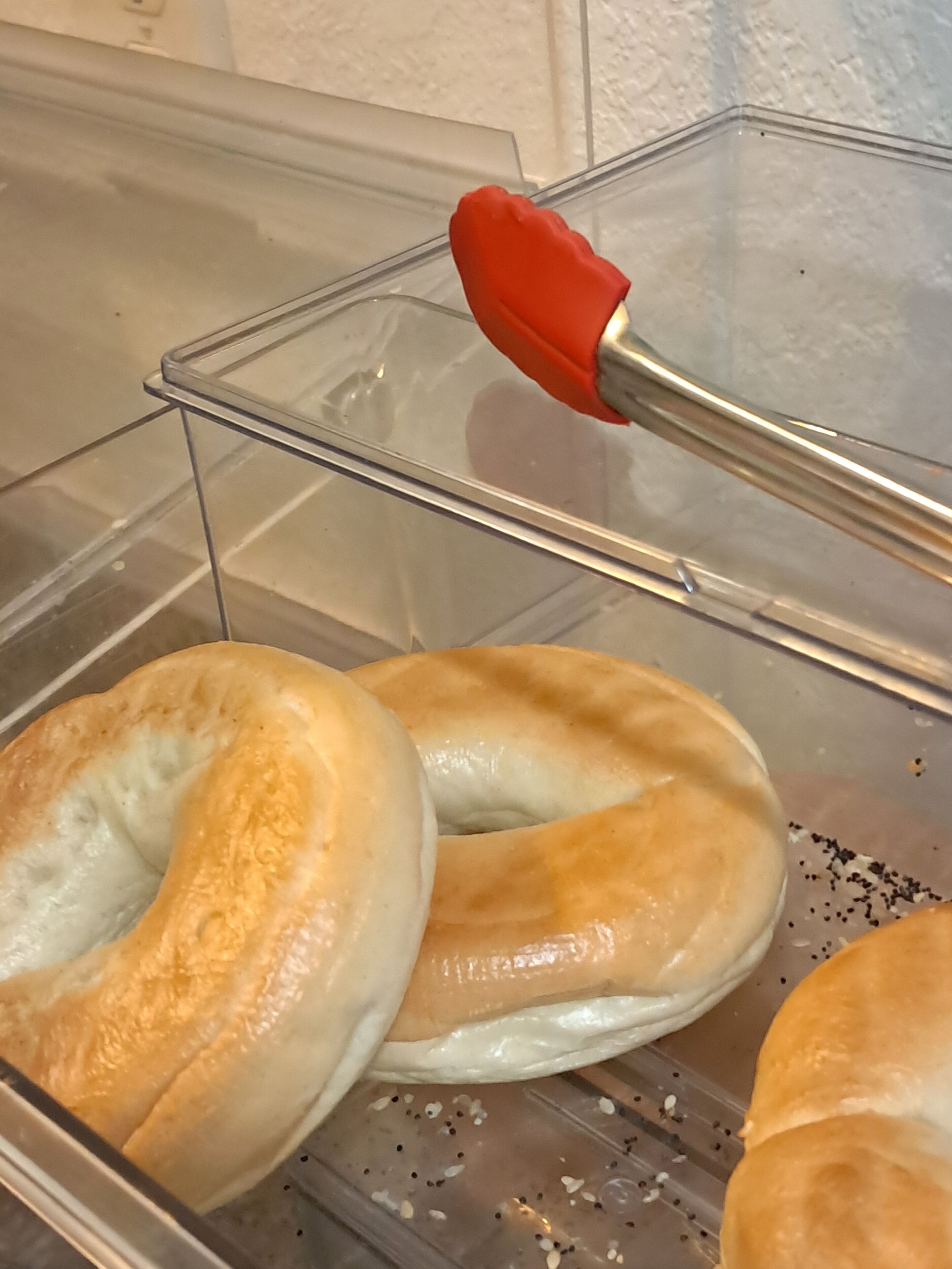 A clear plastic container with a red-handled tool, possibly a spatula, and several bagels with a golden-brown crust.