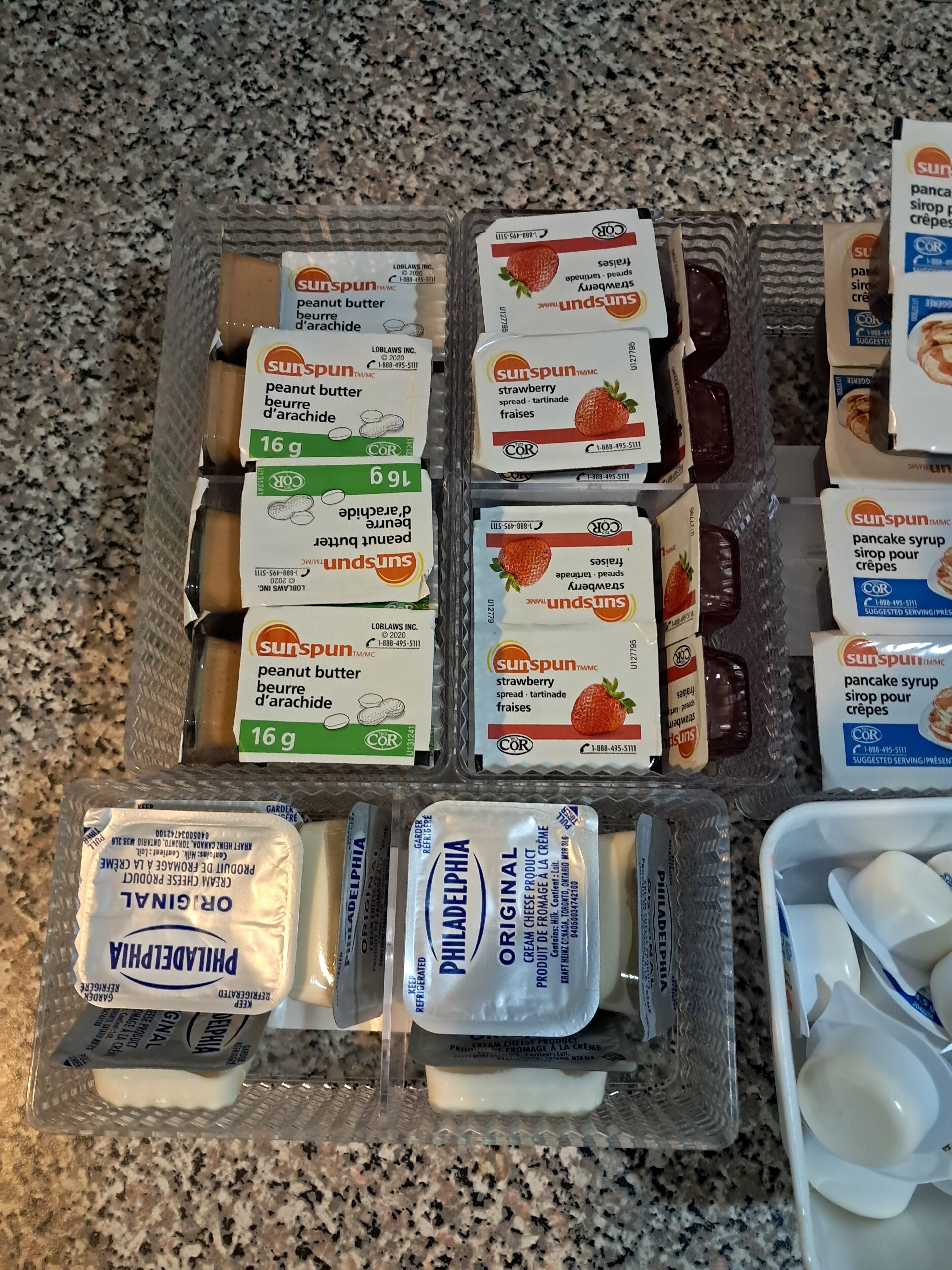 Variedad de alimentos, incluyendo crema de cacahuete, mermelada de fresa, sirope para panqueques y queso crema Philadelphia, todo en un recipiente de plástico.