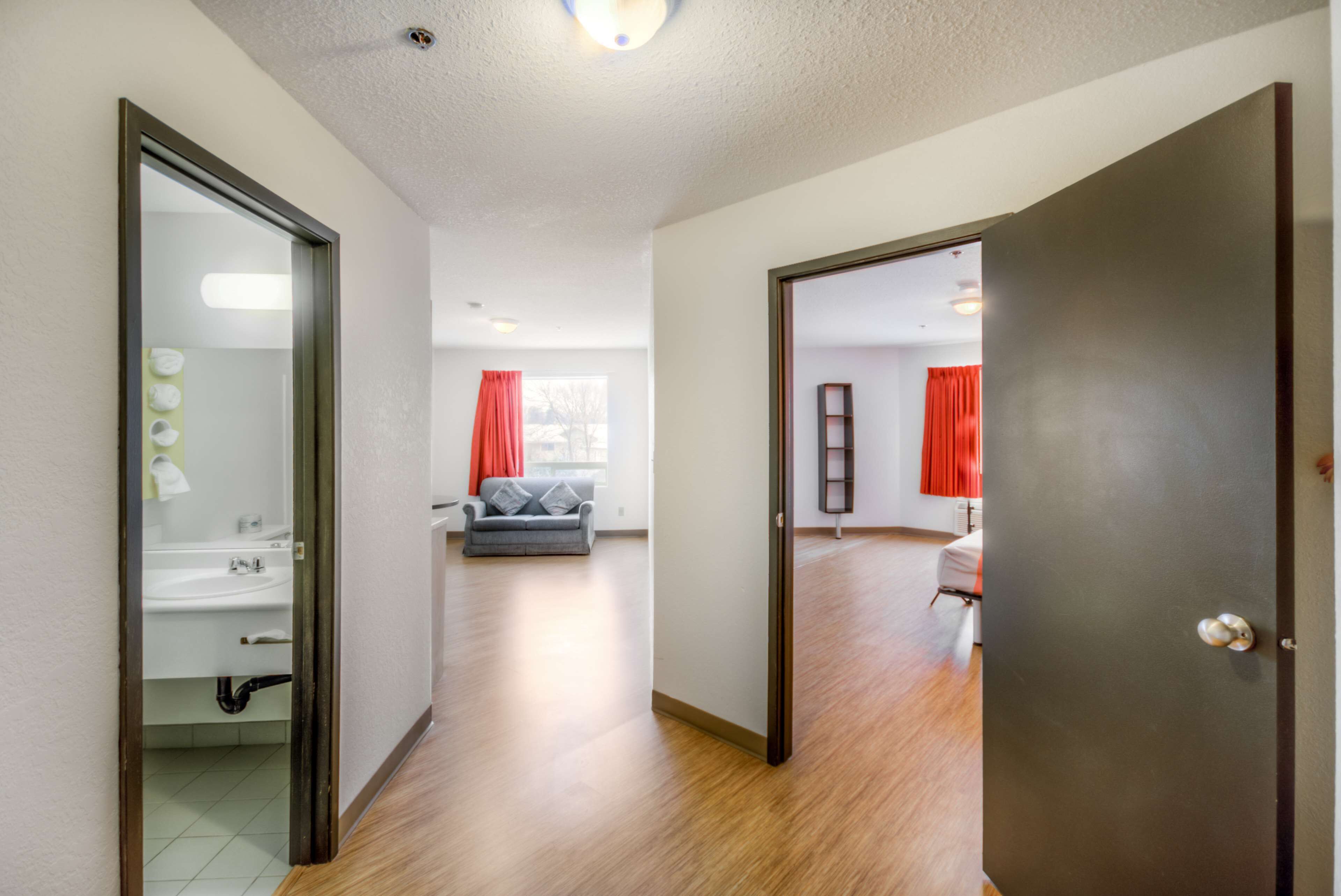 A hallway with a bathroom on the left, a grey sofa in the center, and a door leading to another room on the right.