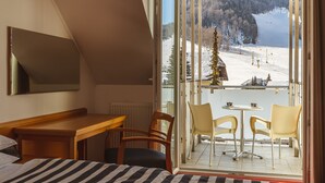 A room with a bed, a desk, and a balcony with chairs and a table. The balcony overlooks a snowy landscape with trees and a ski slope.