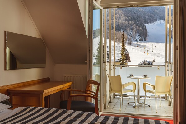 A room with a bed, a desk, and a balcony with chairs and a table. The balcony overlooks a snowy landscape with trees and a ski slope. - Ramada Hotel & Suites by Wyndham Kranjska Gora (Kranjska Gora)