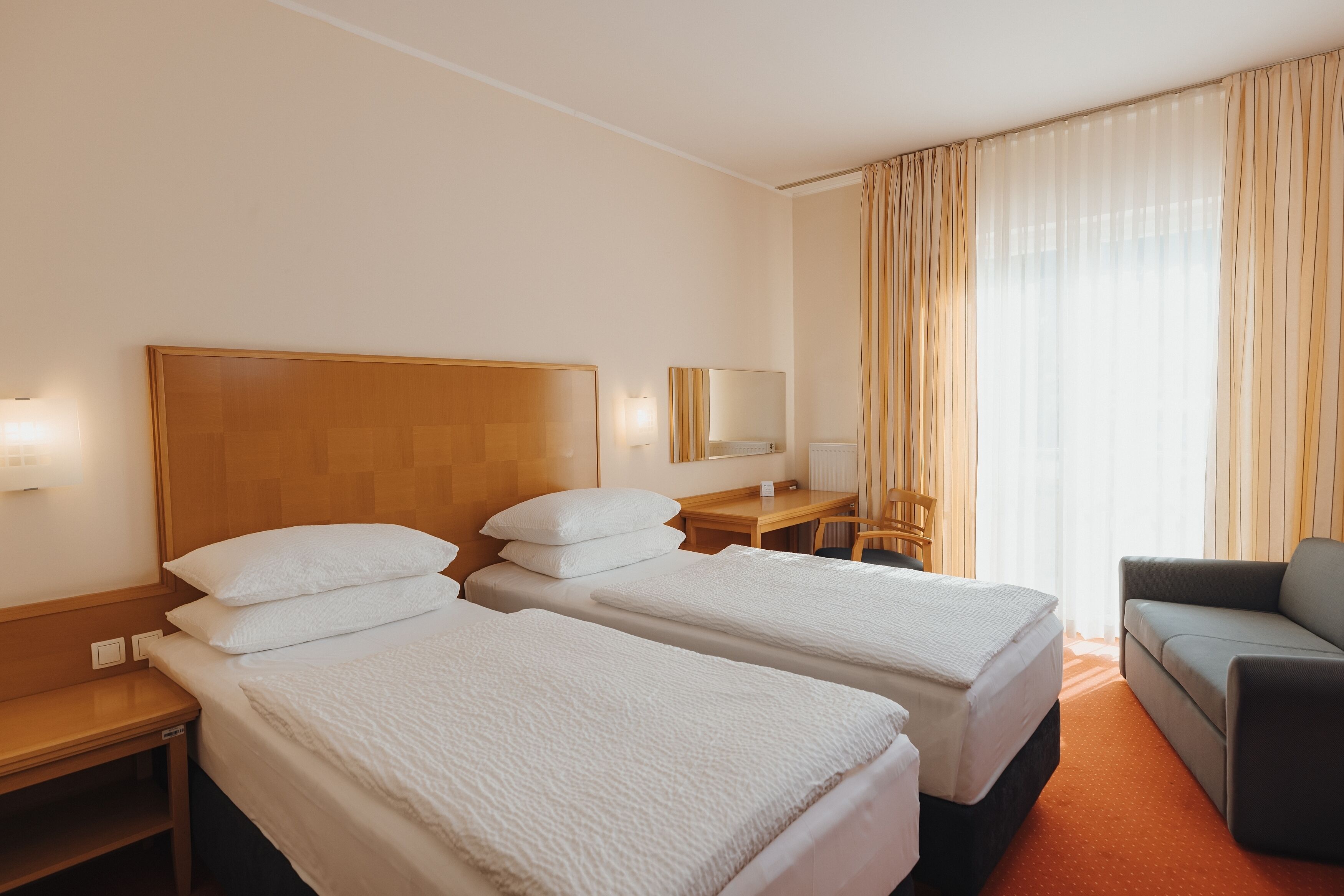 Quarto de hotel com duas camas de solteiro, uma escrivaninha, uma cadeira e um sofá.
