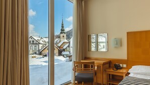 A hotel room with a large window, a desk, a chair, a bed, and a view of a church and snowy street.