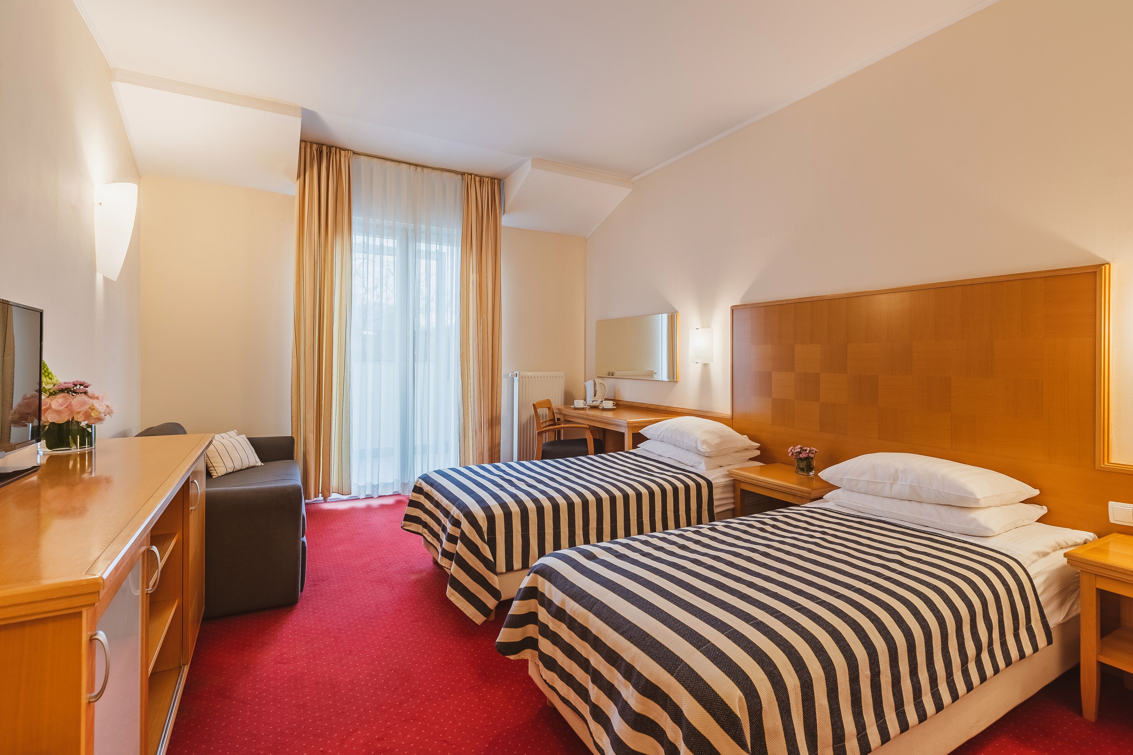 Habitación de hotel con dos camas, un escritorio, una silla, un aparador y un ventanal con cortinas.