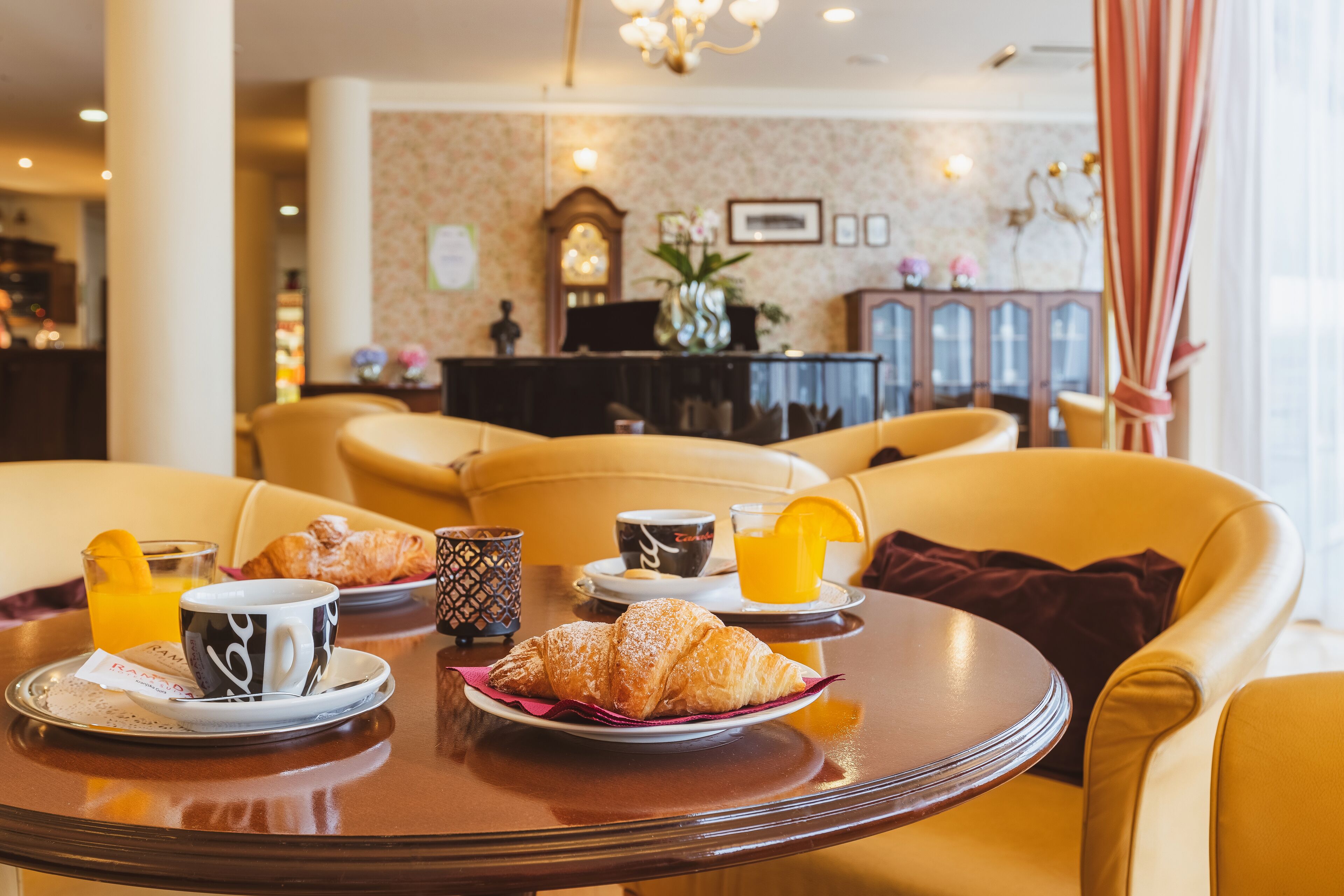 Un desayuno con croissants, café y jugo de naranja sobre una mesa en el vestíbulo de un hotel, con sillas amarillas y un piano al fondo.