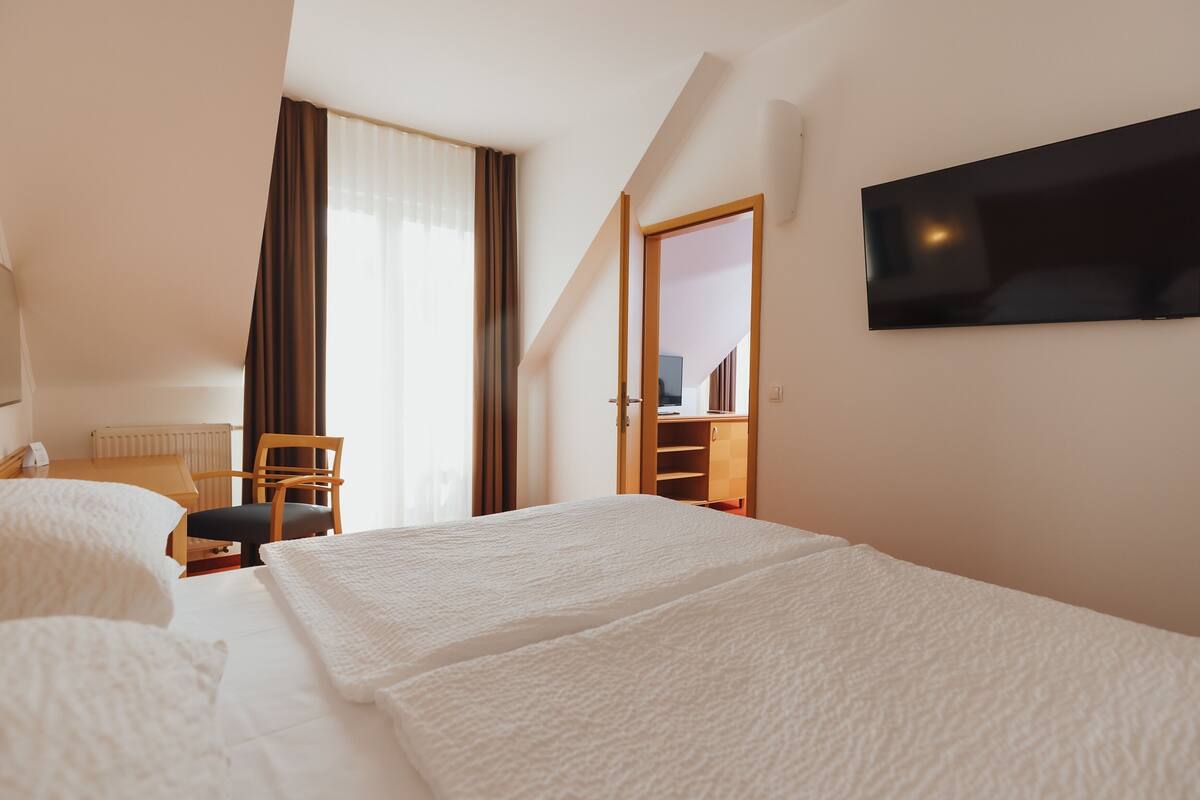 Habitación de hotel con dos camas, un televisor en la pared, un escritorio con silla y una ventana con cortinas.