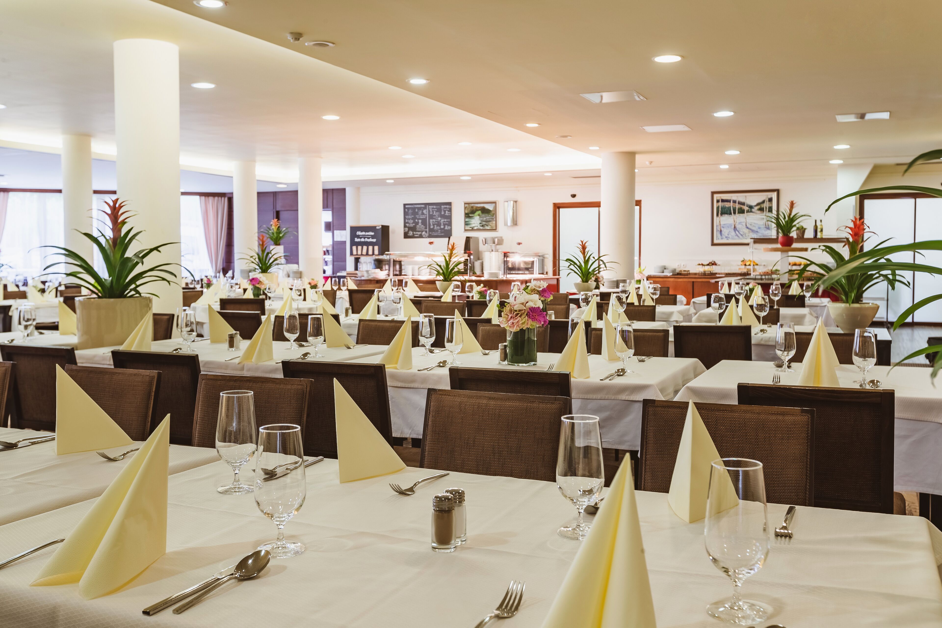 A dining area with tables set for service, featuring white tablecloths, yellow napkins, and clear glassware.