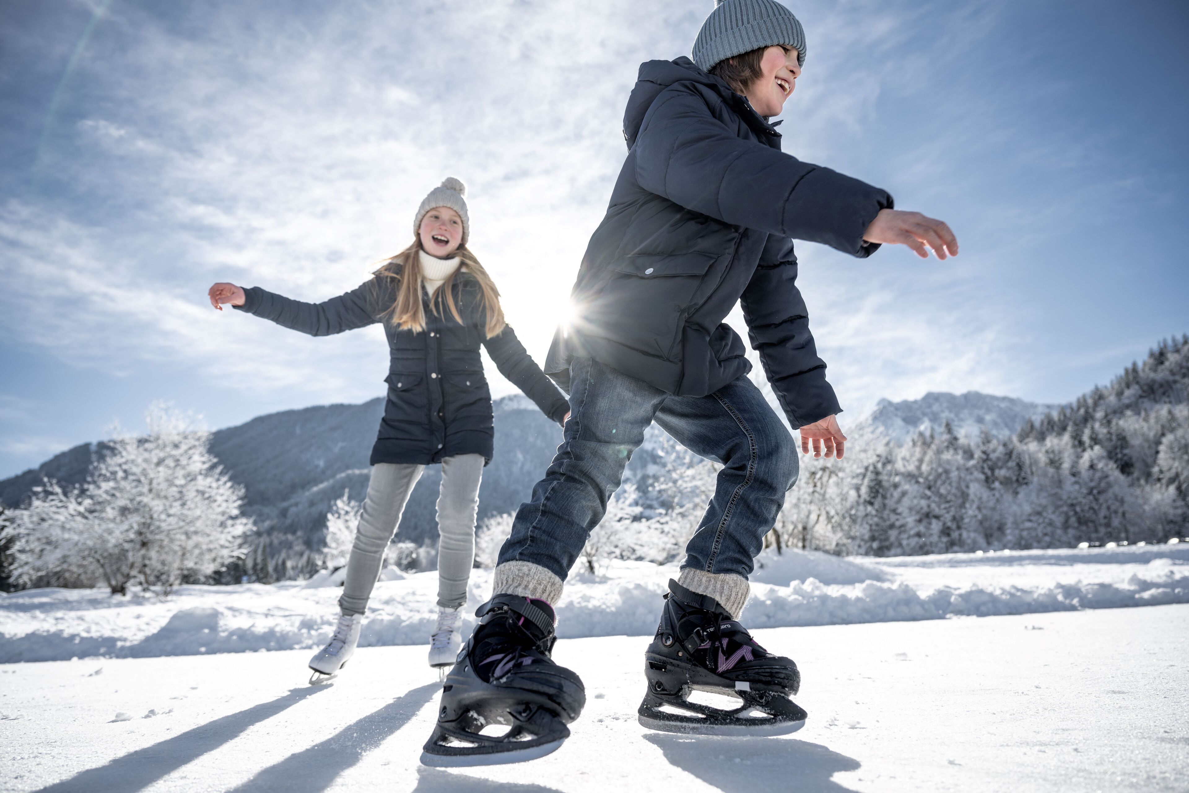 Drei Personen laufen auf einer verschneiten Fläche mit Bergen im Hintergrund Eislaufen.
