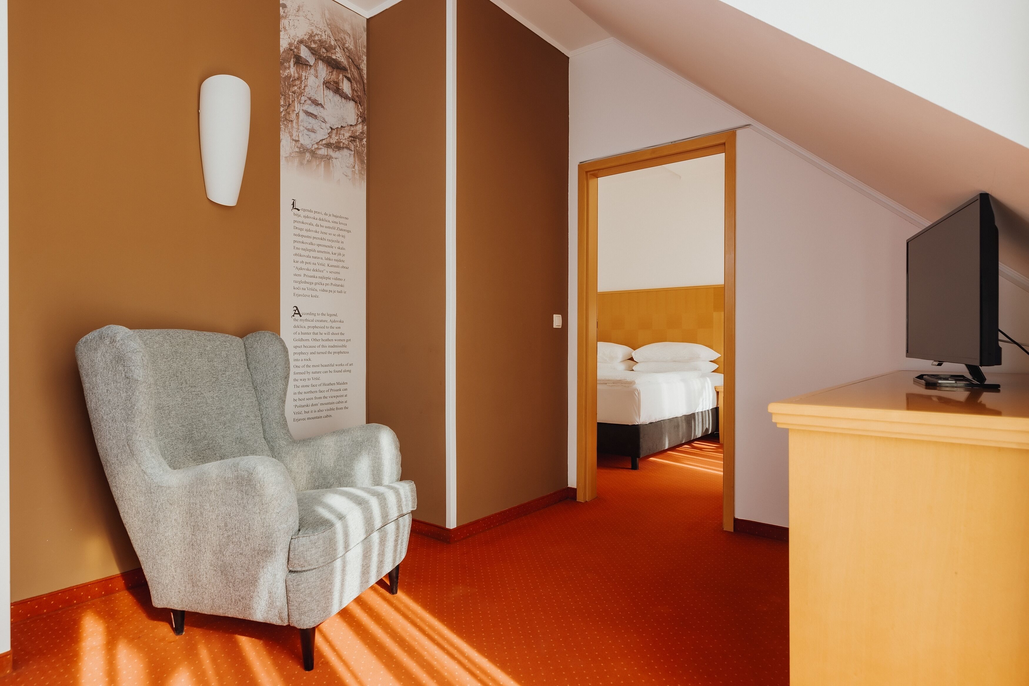 Ein Hotelzimmer mit grauem Sessel, einem Holztisch mit Fernseher und einem Bett mit weißer Bettwäsche.