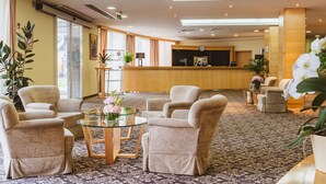 A hotel lobby with a reception desk, seating area with armchairs and a glass table, and decorative plants.