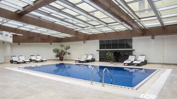 An indoor swimming pool with clear glass ceilings, lounge chairs, and potted plants.