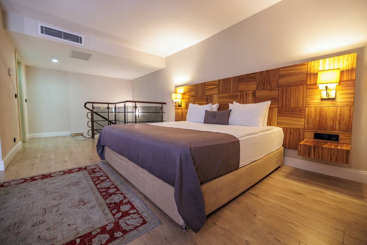 Ein Schlafzimmer mit einem hölzernen Kopfende, einem Bett mit grau-weißer Decke, einem rot-beigen Teppich und einem Nachttisch mit Lampe.