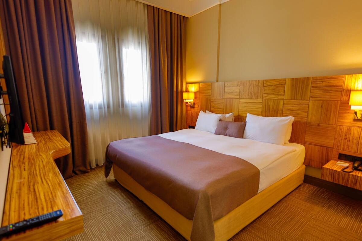 Ein Hotelzimmer mit einem großen Bett, einem Holzkopfende, Nachttischlampen, einem Nachttisch aus Holz und einer Fernbedienung auf einem Holztisch. Ein Fenster mit Vorhängen ist ebenfalls zu sehen.